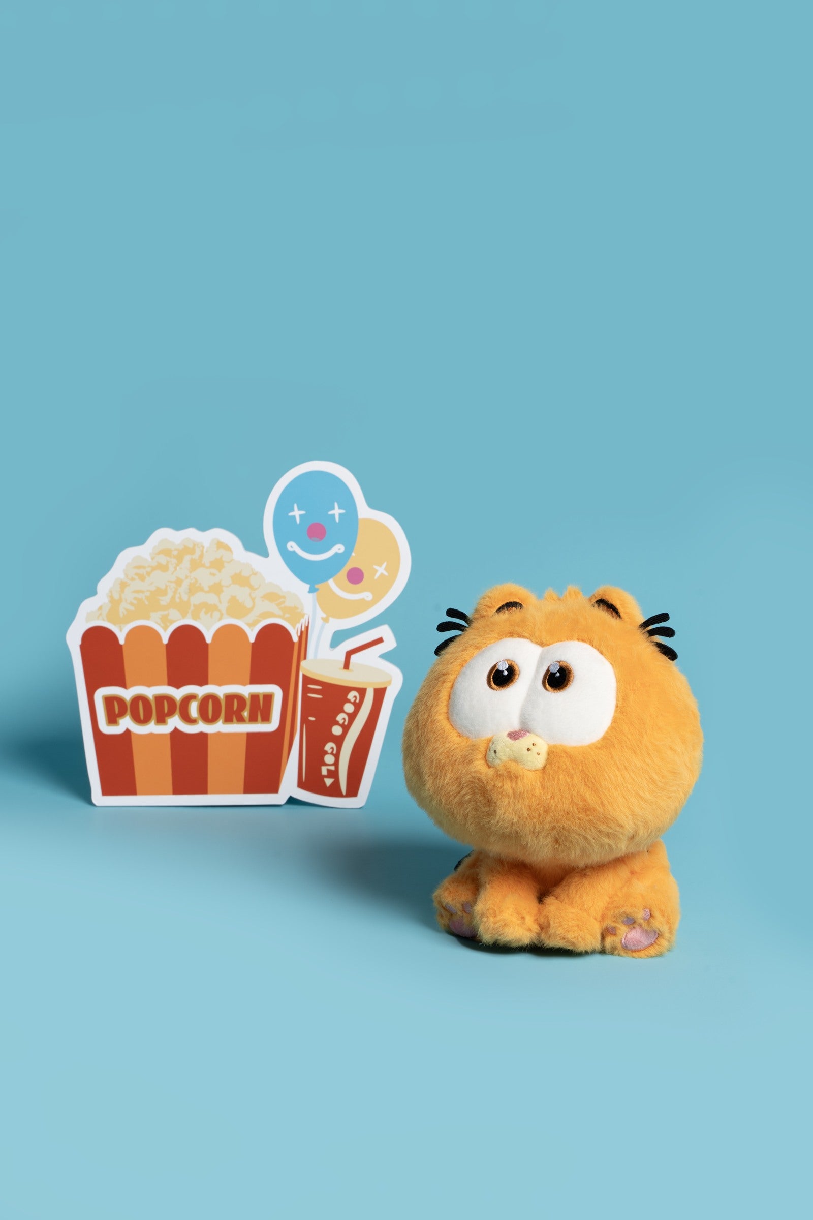 Seated Garfield plush toy beside popcorn and drink illustrations on a blue set