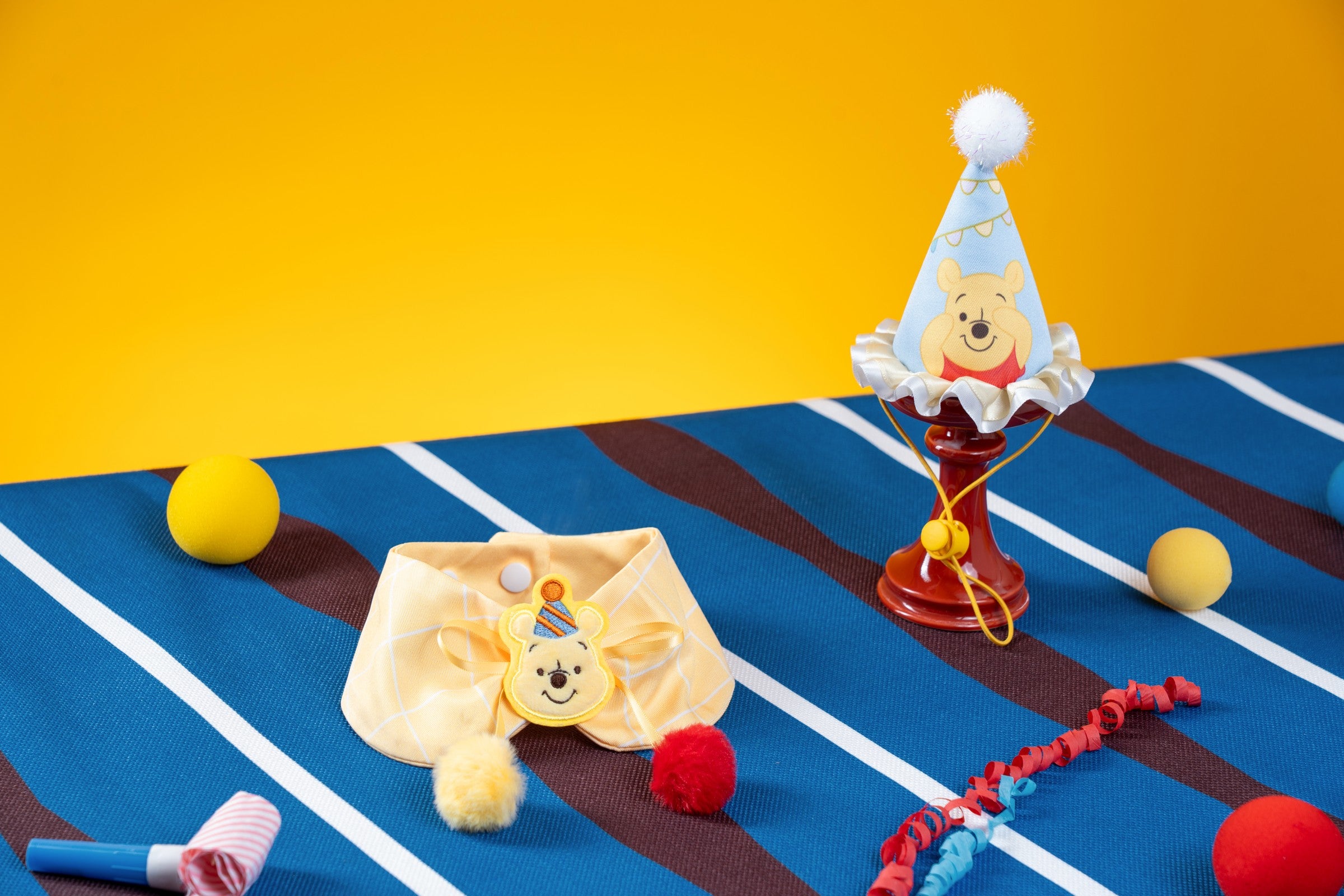 Winnie the Pooh party hat and collar arranged on a blue striped picnic blanket against a yellow backdrop